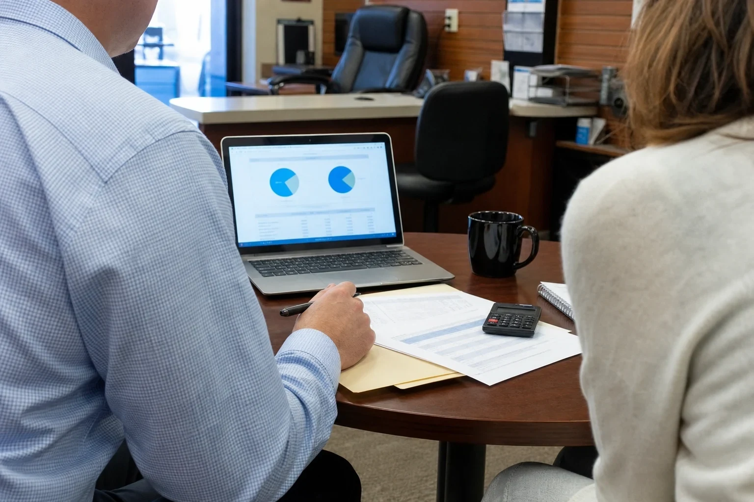 Man in a checked button-down shirt reviewing pie charts on a laptop with a woman at a round wooden table holding papers, a calculator and a black mug in an office